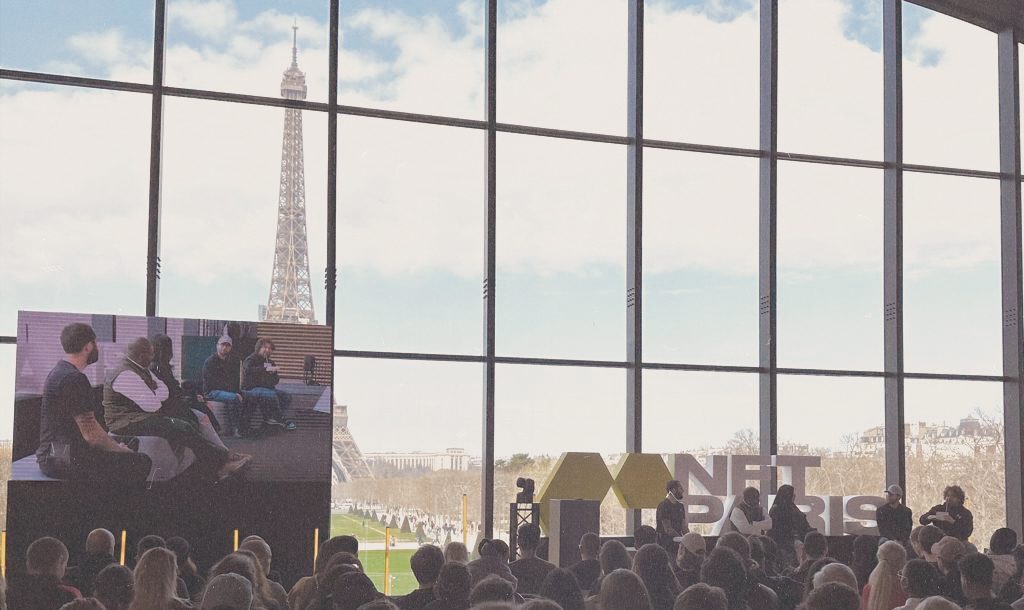 Réunion de personnes lors du salon NFT. Une grande baie vitrée fait face à la Tour Eiffel. 